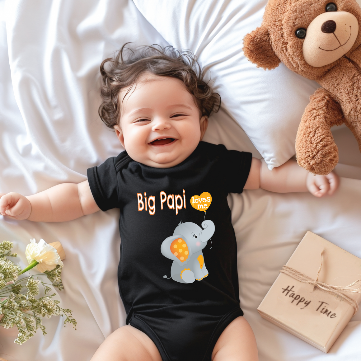 A smiling baby wearing a black t-shirt with an elephant design lies on a bed next to a stuffed teddy bear.