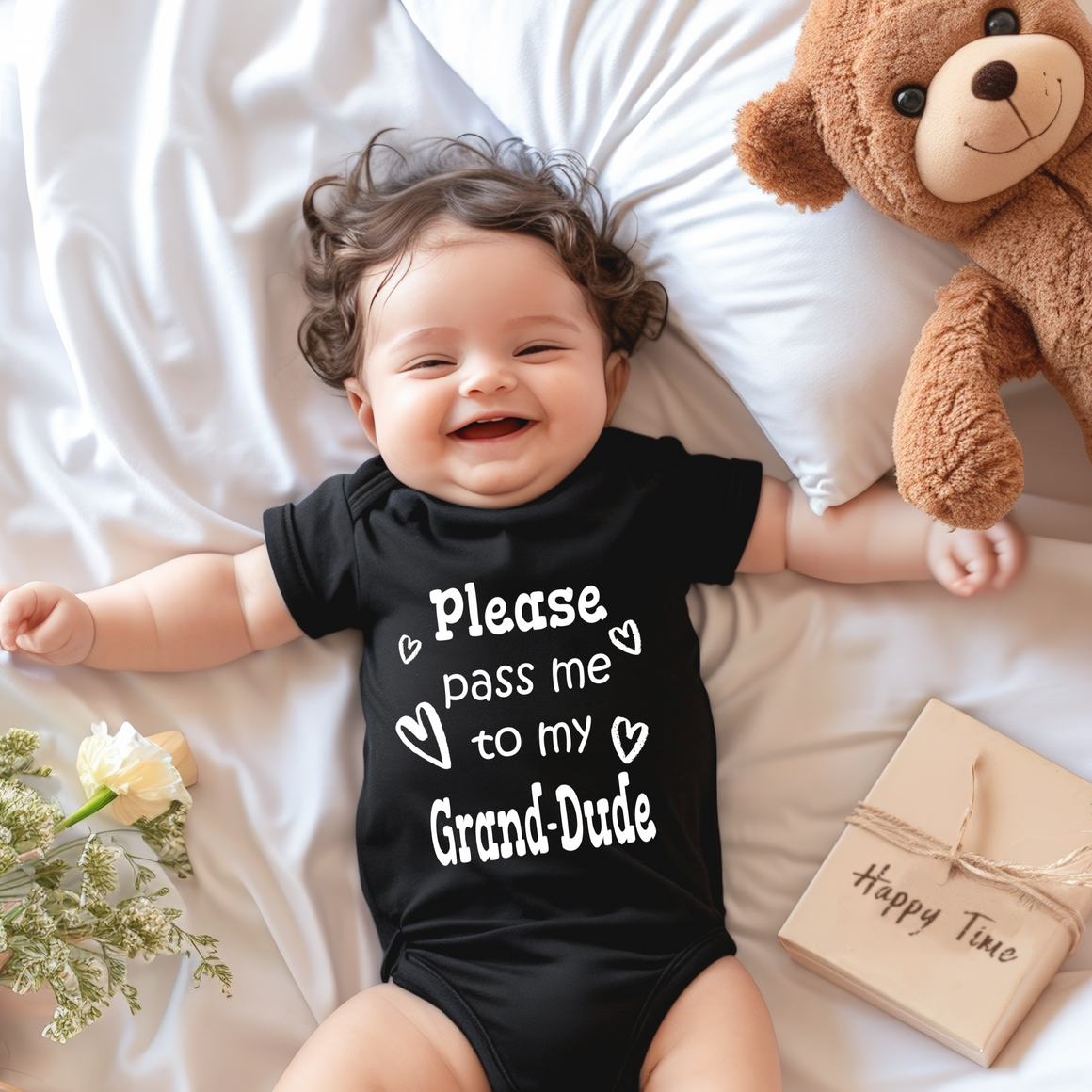 A smiling baby wearing a black onesie with the text "Please pass me to my Grand-Dude" lies on a bed next to a teddy bear and a bouquet of flowers.