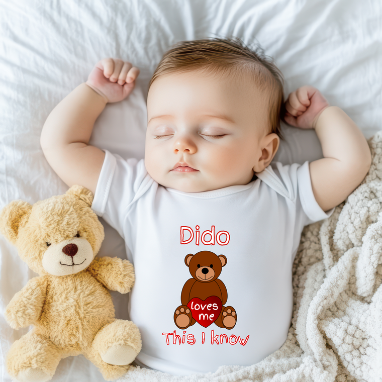 A sleeping baby wearing a white onesie with a teddy bear and text printed on it.