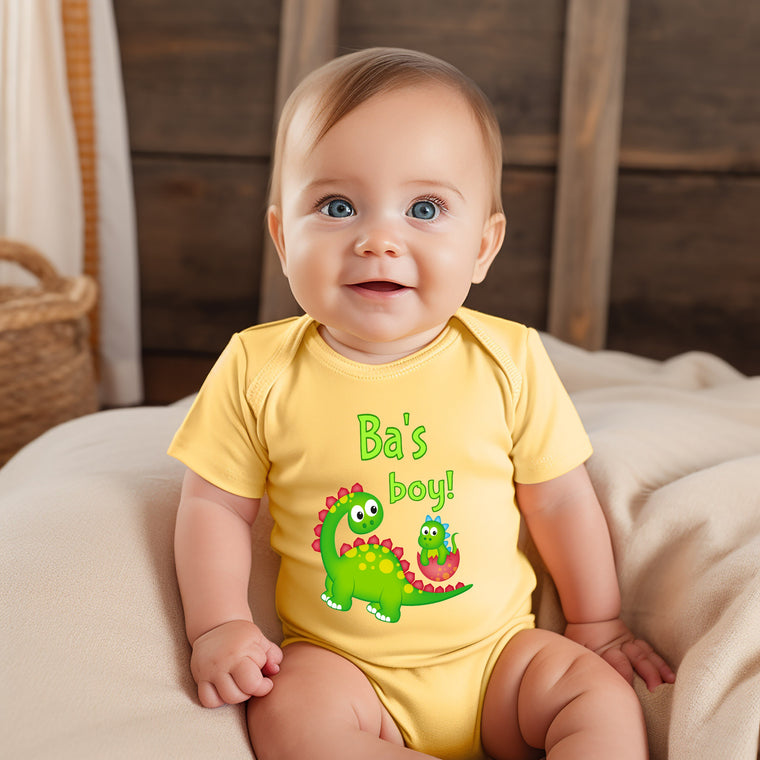 A smiling baby boy wearing a yellow onesie with a dinosaur graphic and the text "Ba's boy!"