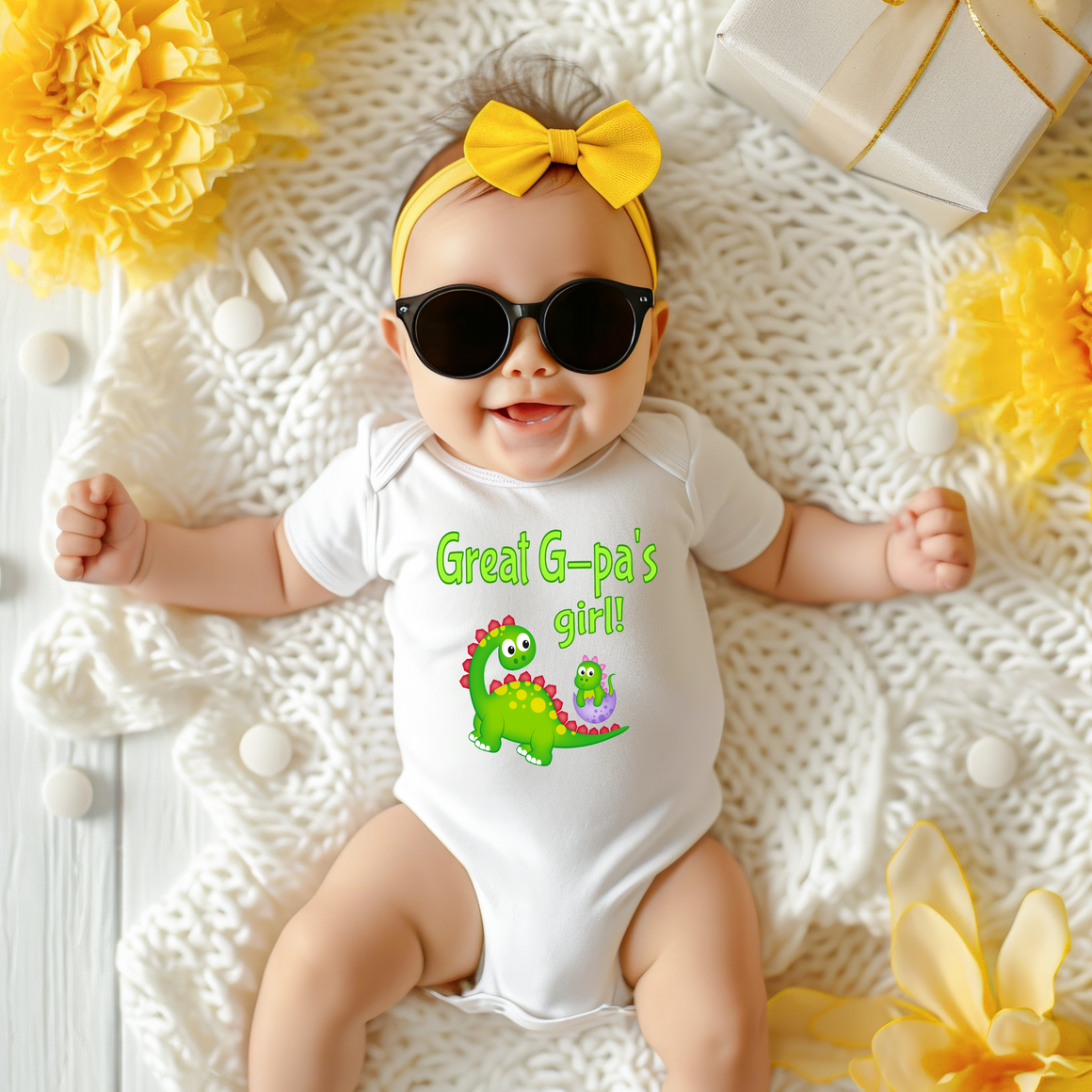 A baby wearing a white onesie with a dinosaur design and sunglasses lying on a white blanket with yellow flowers.