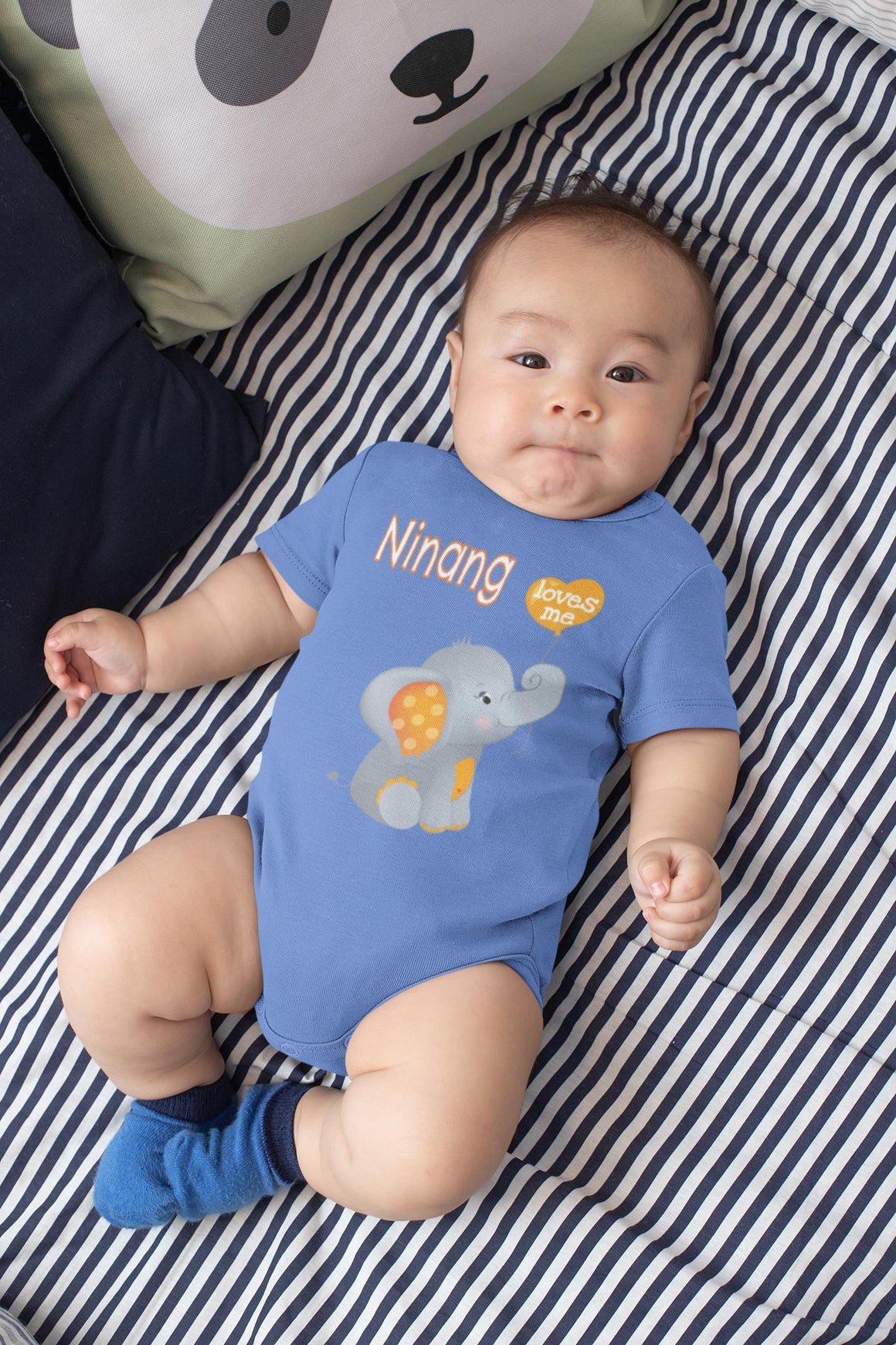 A baby wearing a blue onesie with an elephant design is lying on a striped bed.