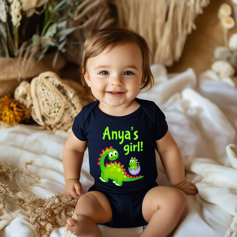A smiling baby girl wearing a black t-shirt with a dinosaur graphic and the text "Anya's girl!"
