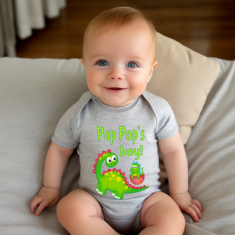 A smiling baby wearing a gray onesie with a dinosaur graphic and the text "Pop Pop's boy!"