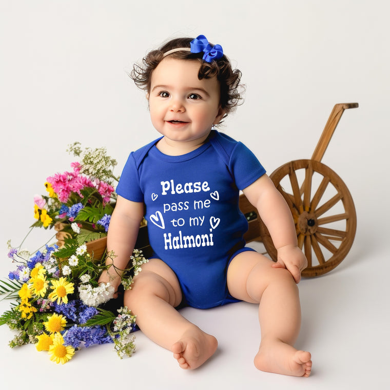 A smiling baby girl wearing a blue onesie with the text "Please pass me to my Halmoni" sits next to a wooden wagon wheel and a basket filled with colorful flowers.
