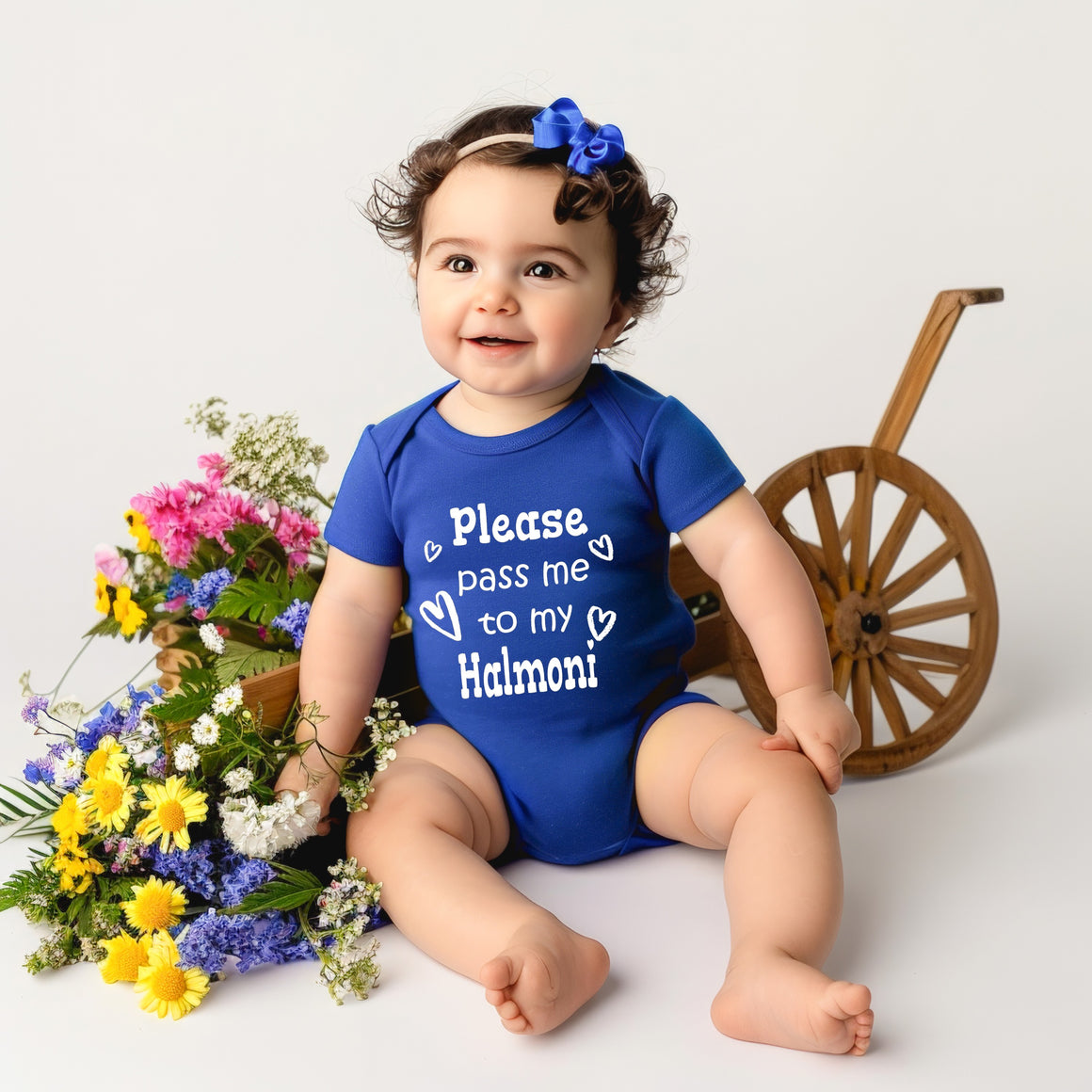 A smiling baby girl wearing a blue onesie with the text "Please pass me to my Halmoni" sits next to a wooden wagon wheel and a basket filled with colorful flowers.