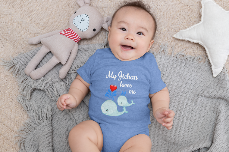 A smiling baby wearing a blue onesie with a whale design lies on a gray blanket, accompanied by a stuffed animal.