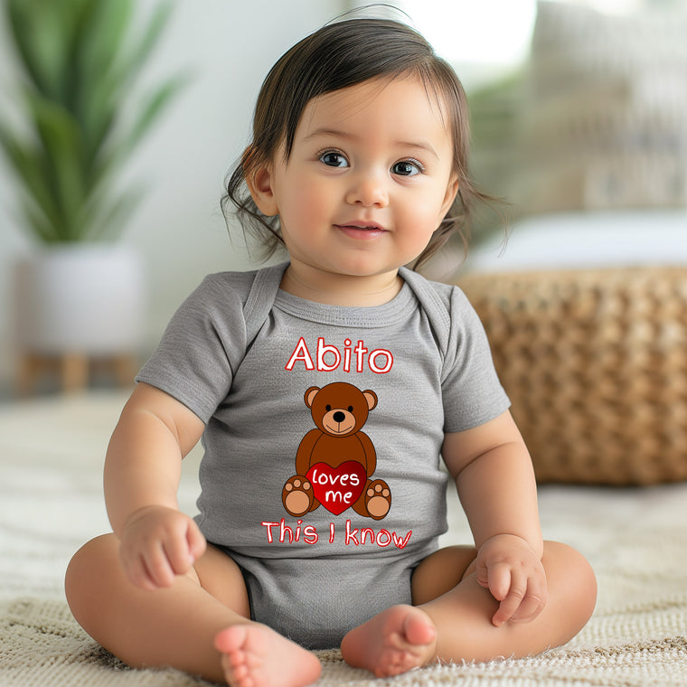 A smiling baby girl wearing a gray onesie with a teddy bear graphic and text.