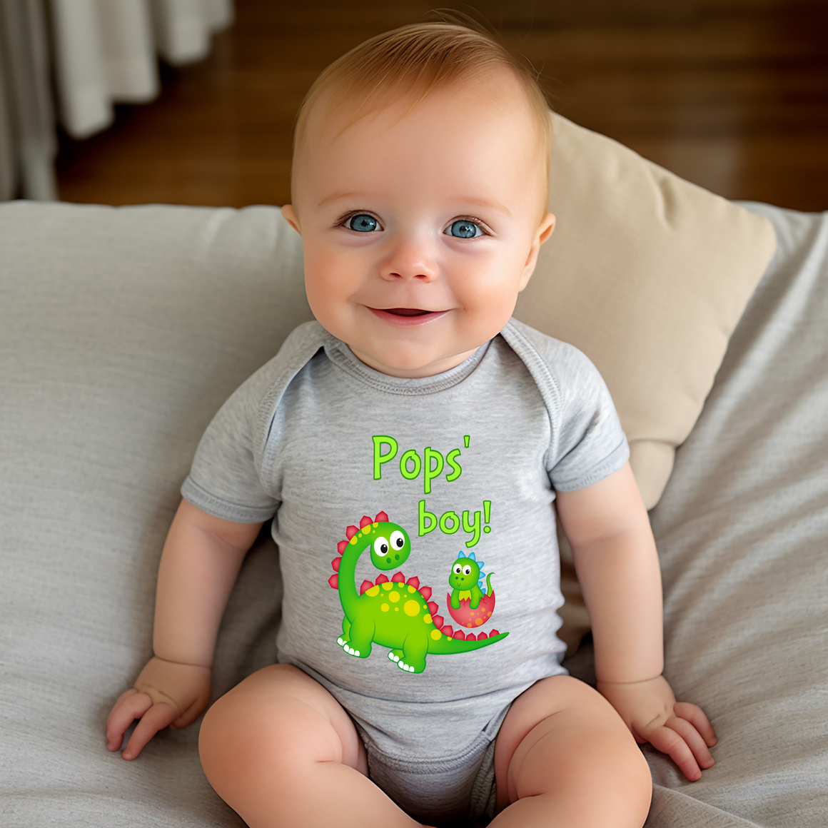 A smiling baby wearing a gray onesie with a dinosaur graphic and the text "Pops' boy!"