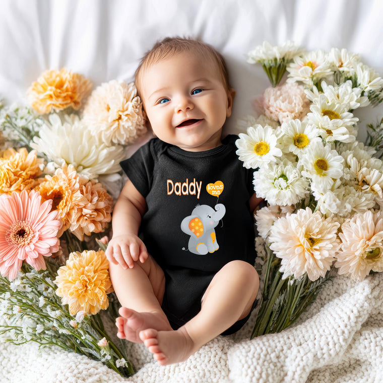 A smiling baby wearing a black onesie with an elephant design lies on a bed surrounded by colorful flowers.