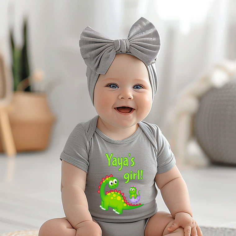 A smiling baby girl wearing a gray headband and a gray t-shirt with a dinosaur graphic, sitting on a white surface.