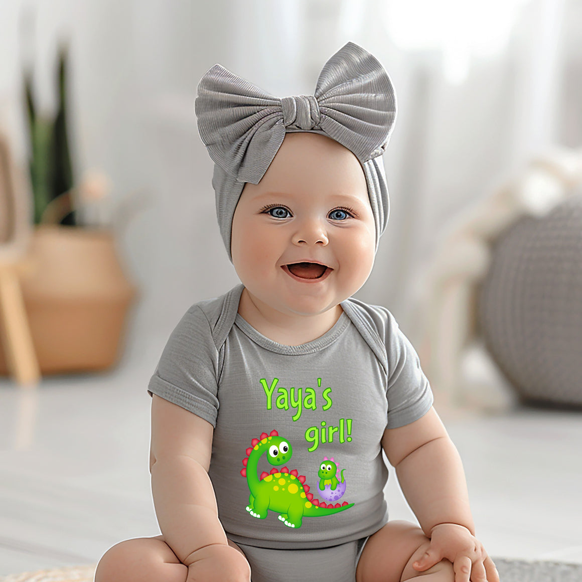 A smiling baby girl wearing a gray headband and a gray t-shirt with a dinosaur graphic, sitting on a white surface.