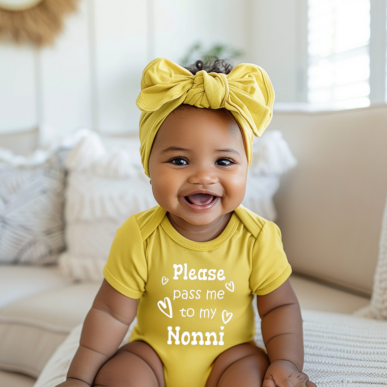 A smiling baby girl wearing a yellow outfit with a bow on her head and a yellow shirt with the text "Please pass me to my Nonna" printed on it.