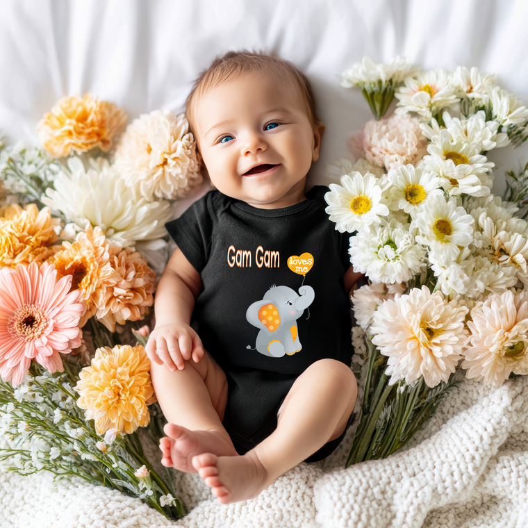 A smiling baby wearing a black outfit with a cartoon elephant design lies on a bed surrounded by colorful flowers.