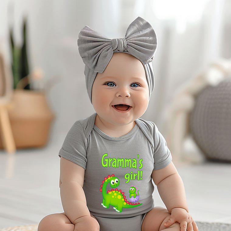 A smiling baby girl wearing a gray headband and a gray onesie with a dinosaur graphic, sitting on a white surface.