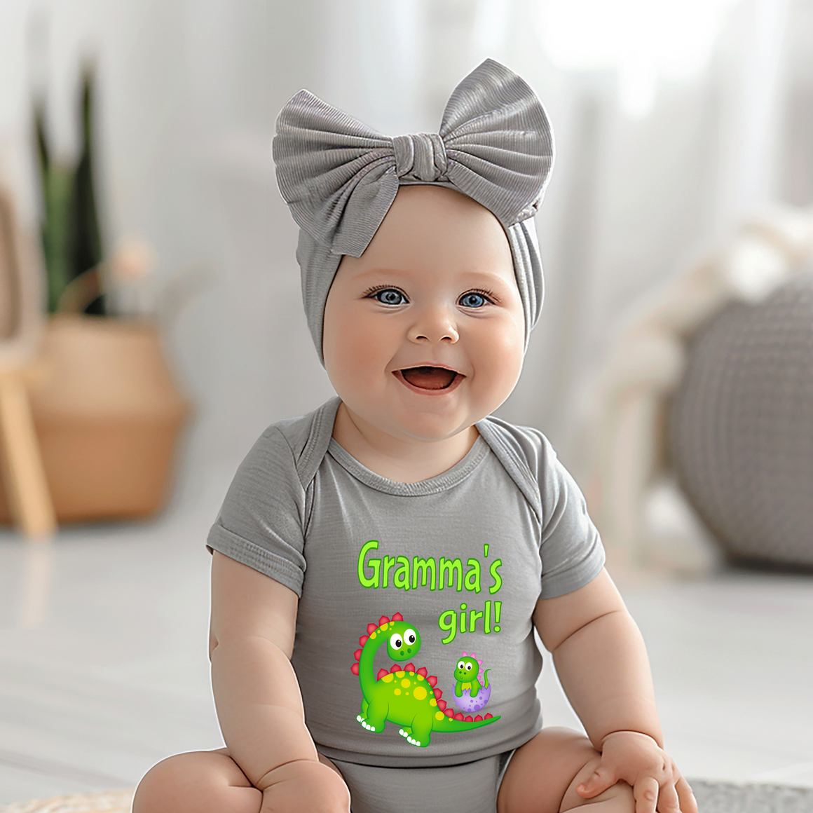 A smiling baby girl wearing a gray headband and a gray onesie with a dinosaur graphic, sitting on a white surface.
