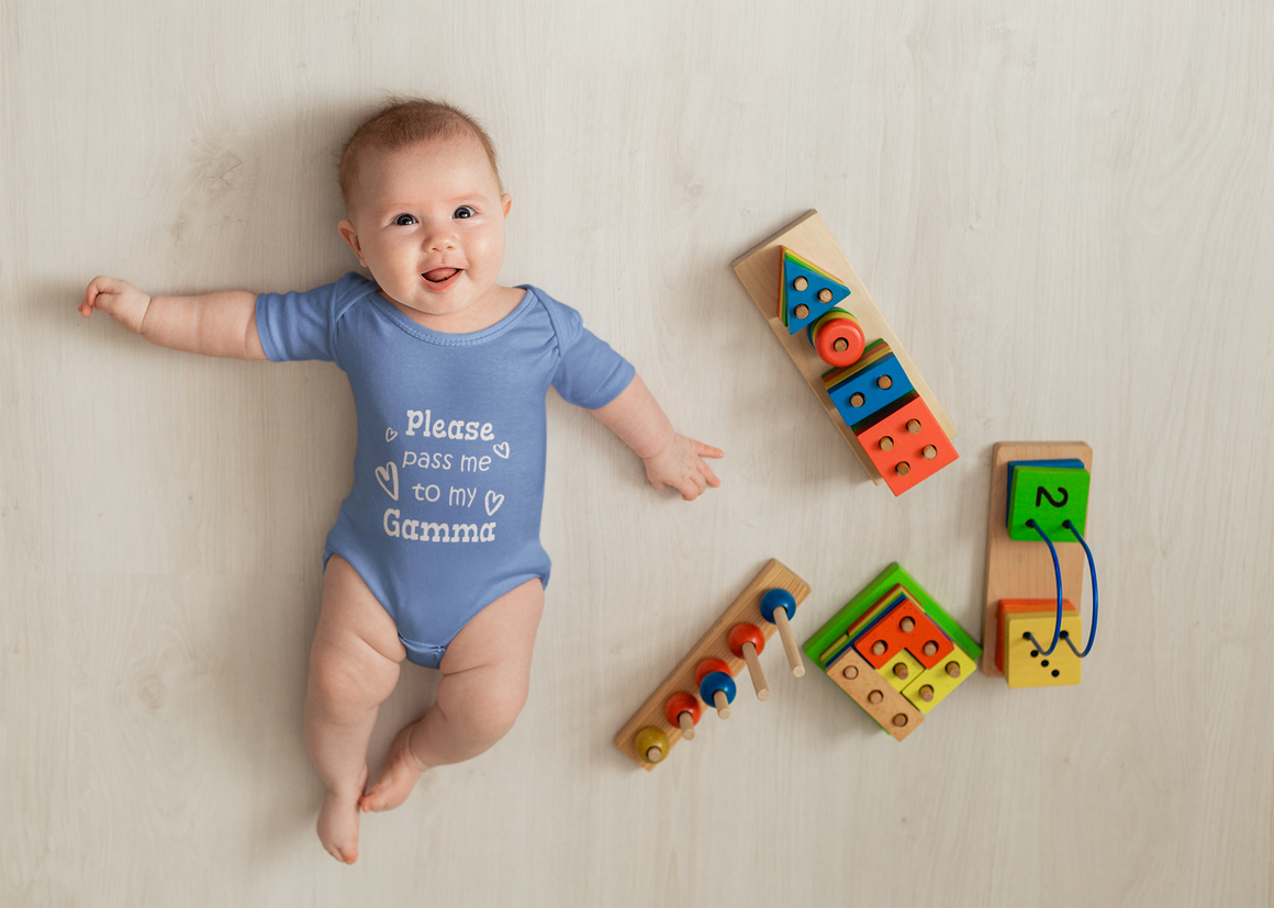 A smiling baby in a blue onesie lying on a wooden floor next to colorful wooden toys.