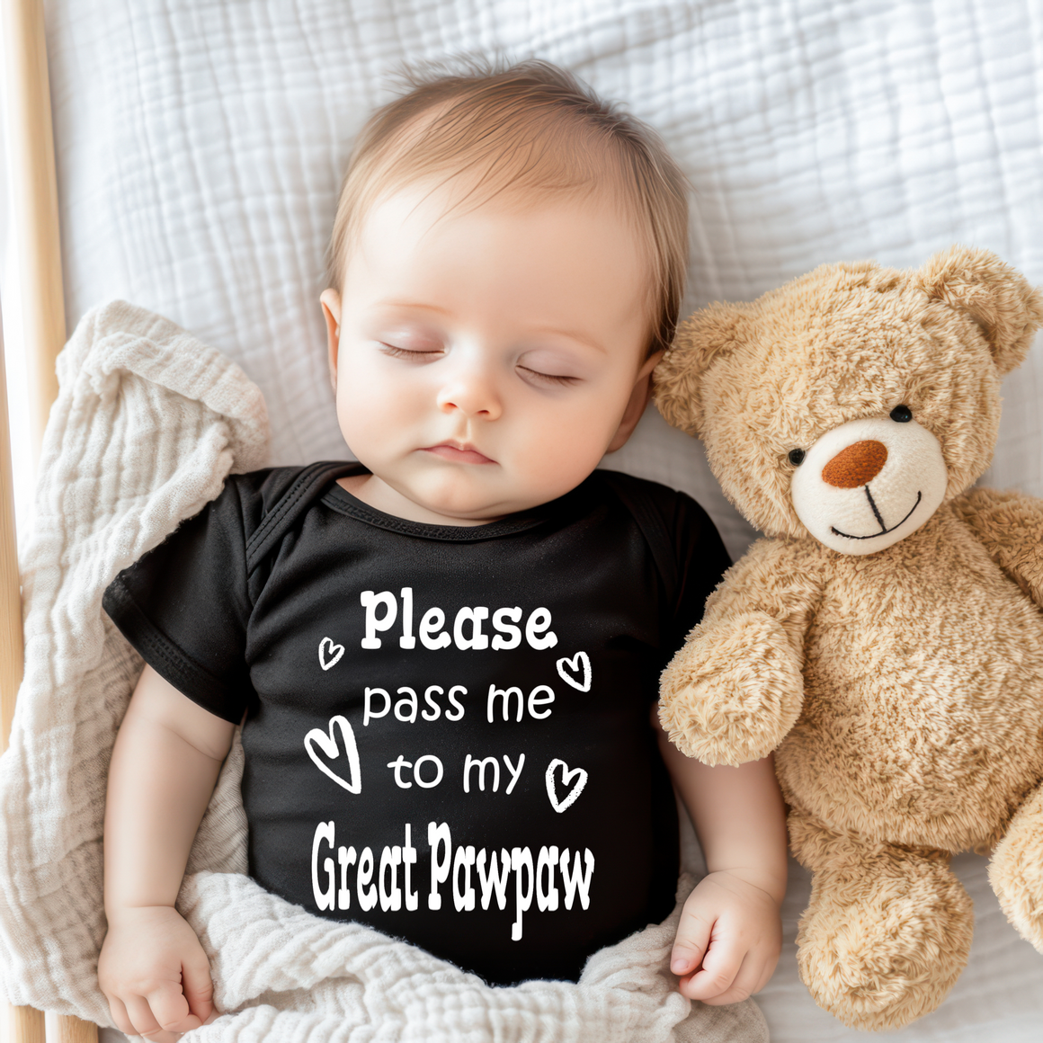 A sleeping baby wearing a black shirt with the text "Please pass me to my Great Paw paw" next to a teddy bear.