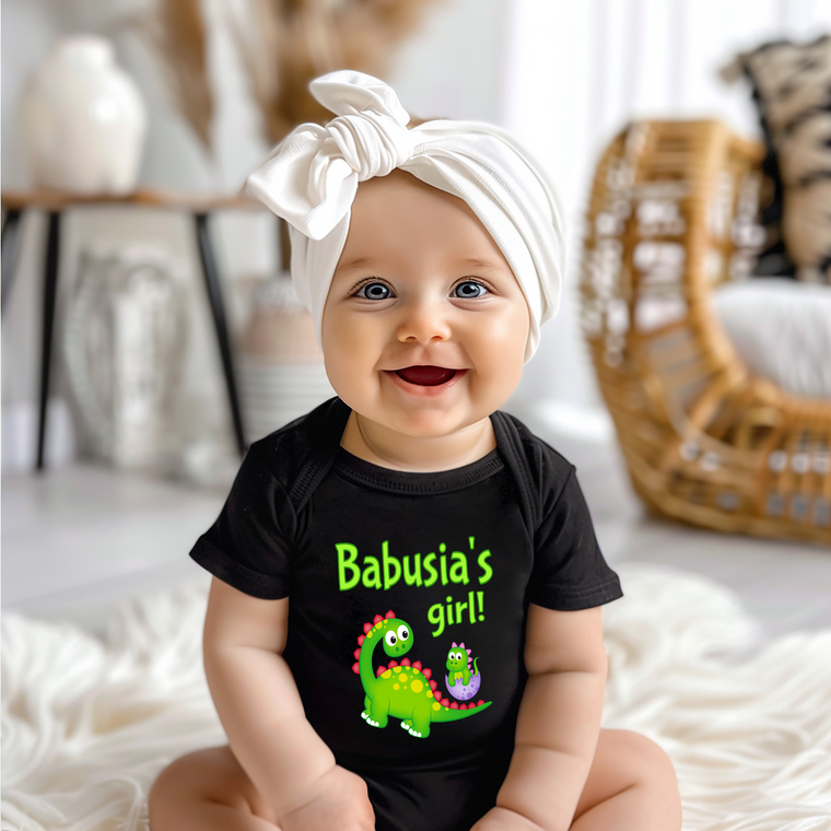 A smiling baby girl wearing a black t-shirt with a dinosaur graphic and the text "Babusia's girl!" is sitting on a white surface.