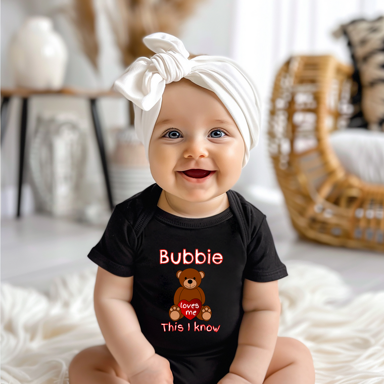 A smiling baby girl wearing a black t-shirt with a teddy bear graphic and the text "Bubbie loves me This I know" is sitting on a white blanket.