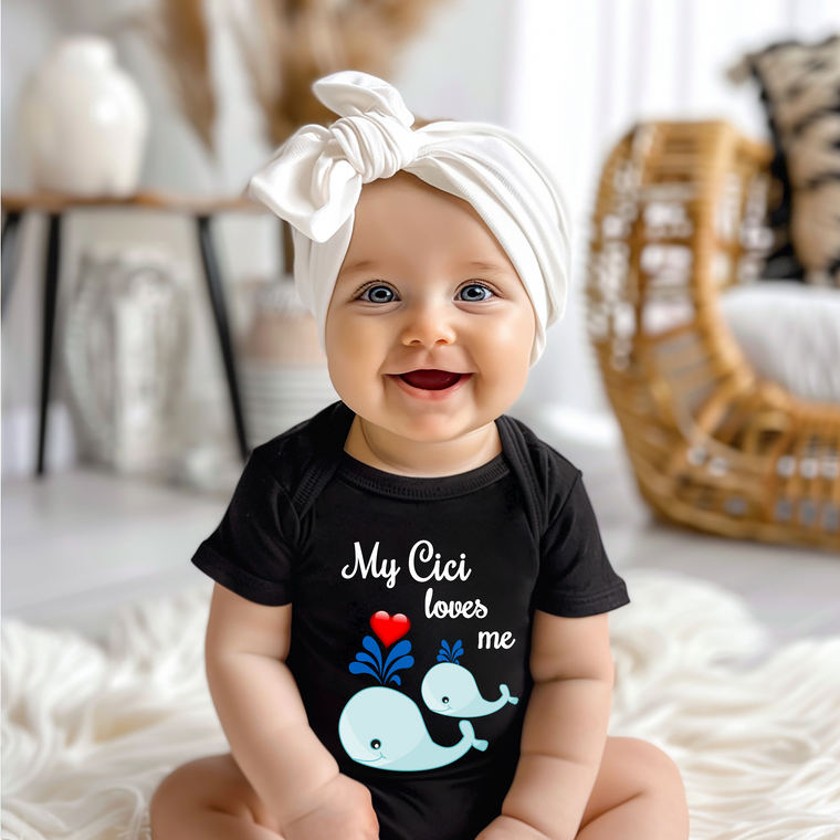 A smiling baby girl wearing a black t-shirt with a whale graphic and the text "My Cici loves me" sits on a white blanket.