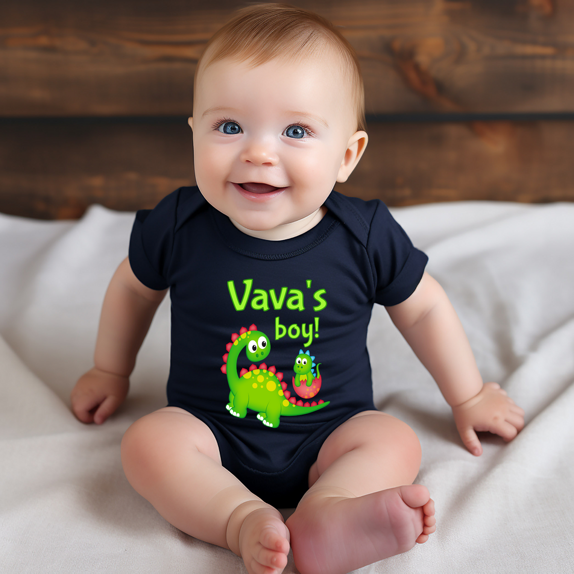 A smiling baby boy wearing a black t-shirt with a dinosaur graphic and the text "Vava's boy!"