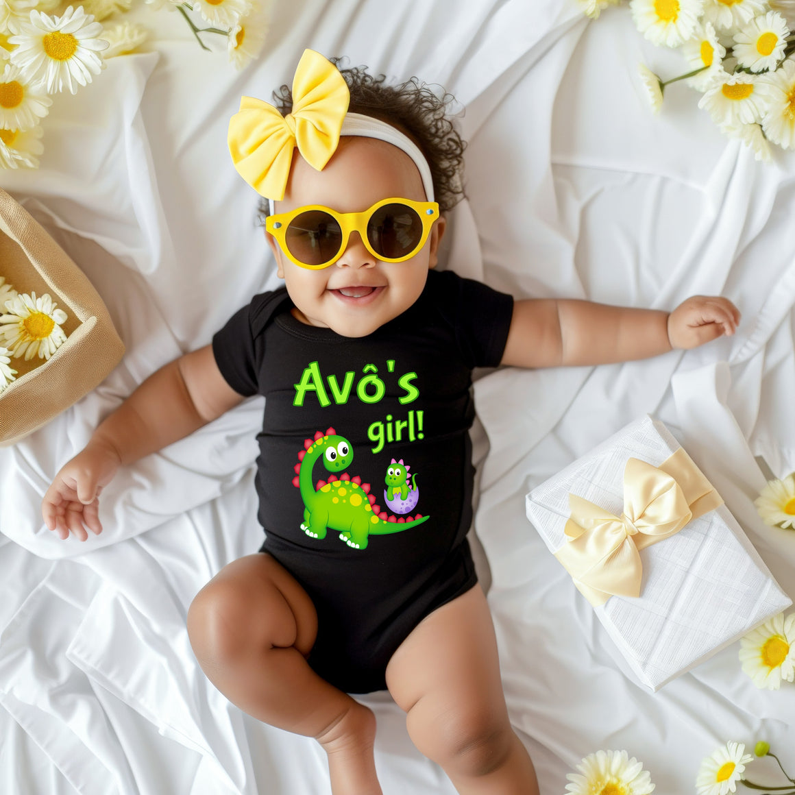 A smiling baby girl wearing a black outfit with a dinosaur and yellow bow sits on a white sheet, surrounded by yellow flowers.