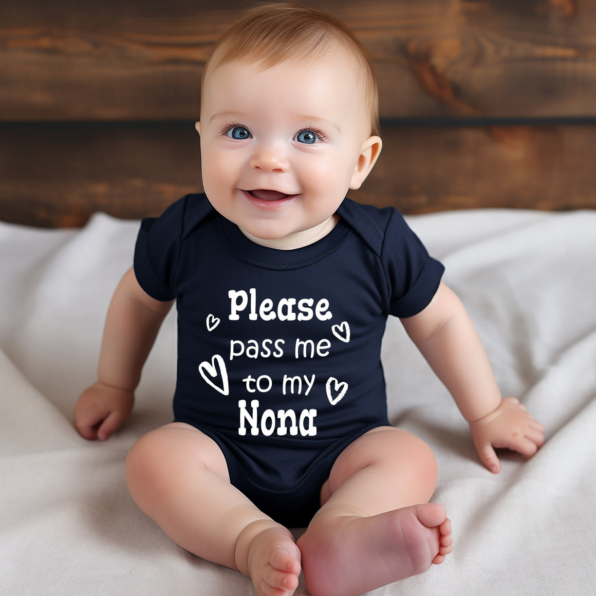 A smiling baby wearing a navy blue onesie with the text "Please pass me to my Nona" printed on it.