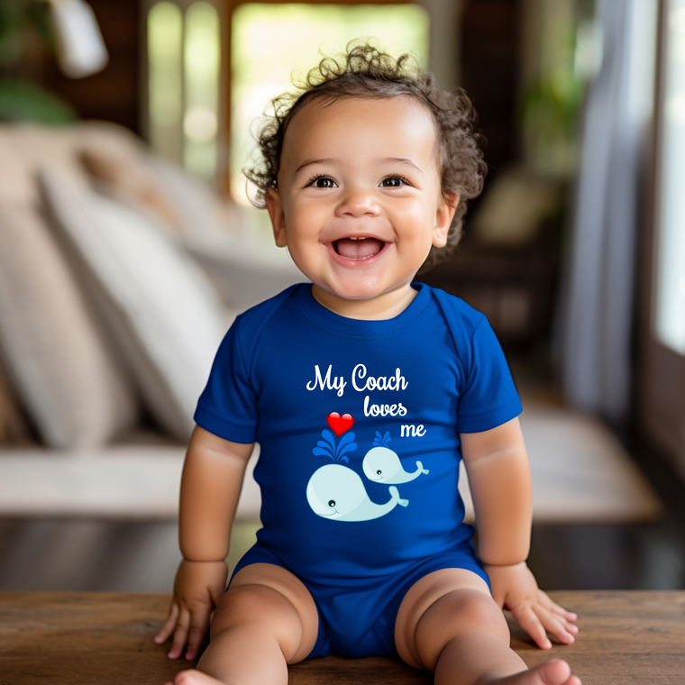 A smiling baby boy wearing a blue shirt with a cartoon whale and the text "My Coach loves me" is sitting on a wooden surface.