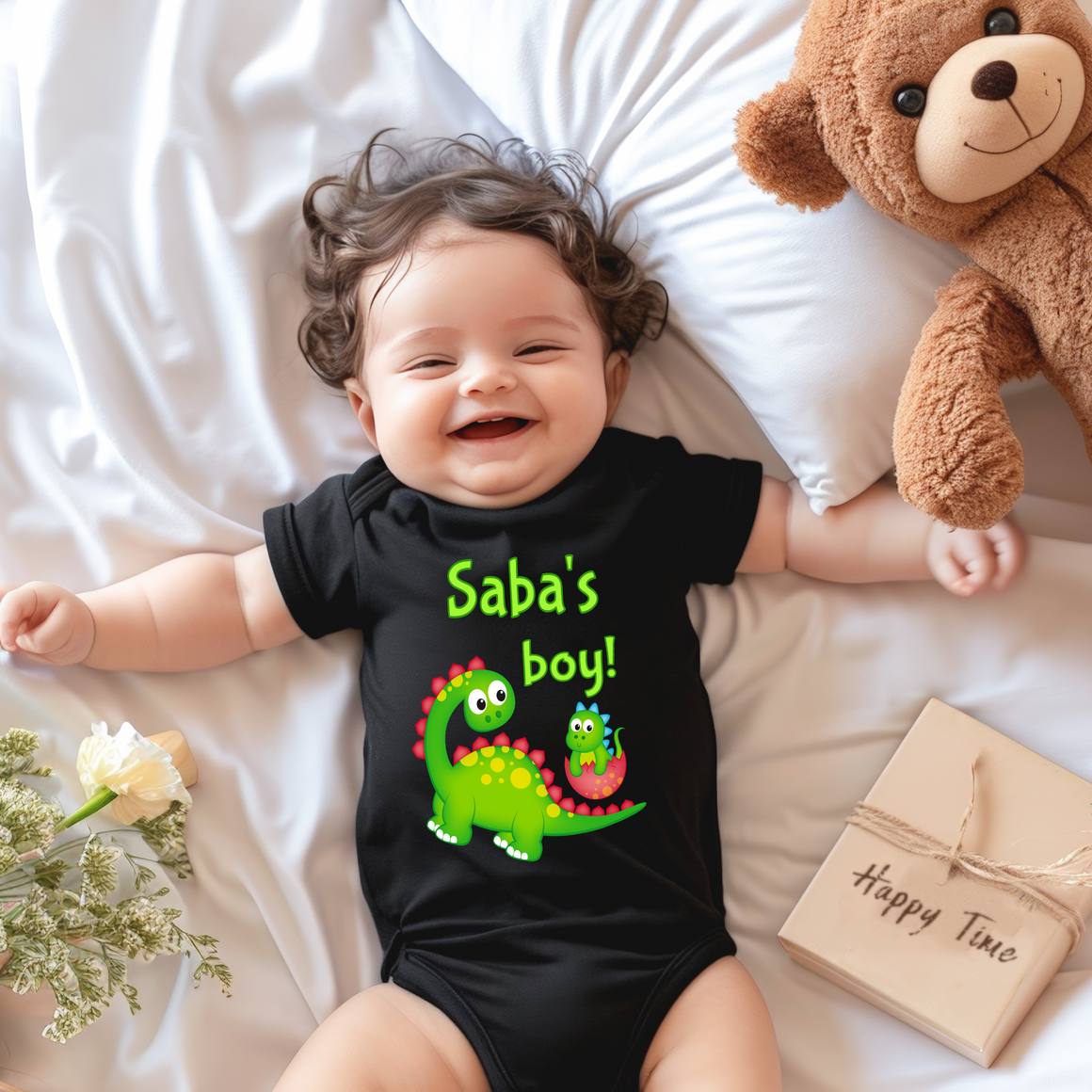 A smiling baby wearing a black t-shirt with a dinosaur graphic lies on a bed next to a stuffed teddy bear.