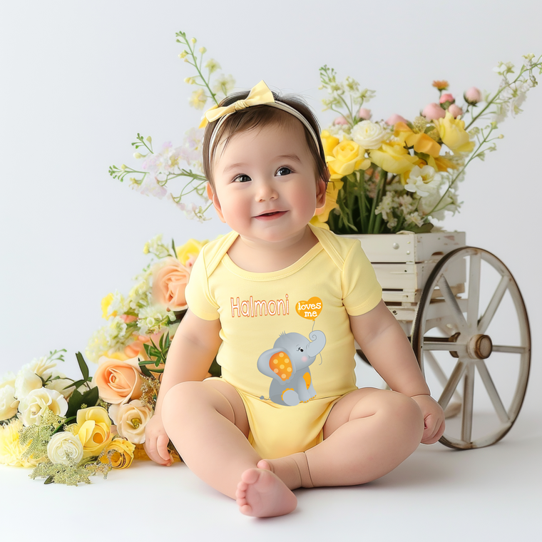 A smiling baby girl wearing a yellow onesie with an elephant design sits in front of a wooden wagon filled with yellow flowers.