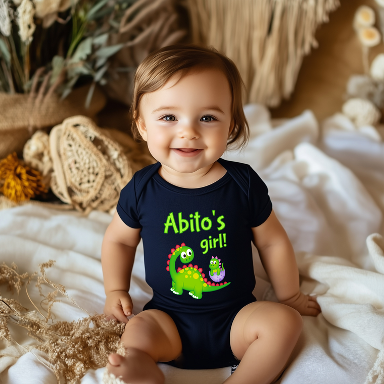 A smiling baby boy wearing a black shirt with a dinosaur graphic and text, sitting on a bed with a floral background.
