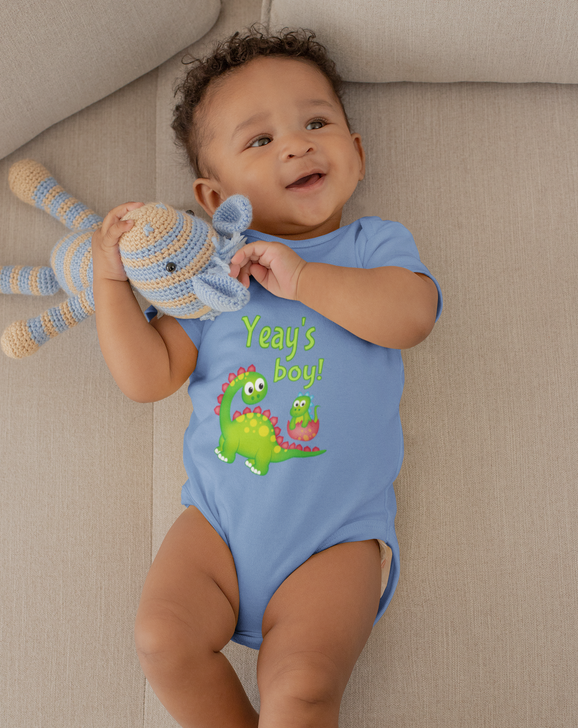 A smiling baby boy wearing a blue onesie with a cartoon dinosaur and the text "Yee's boy!" is lying on a couch.