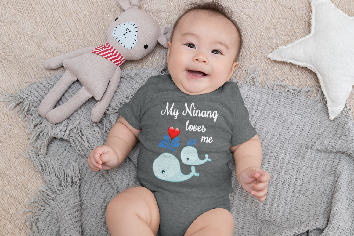 A smiling baby wearing a gray onesie with a design of two blue whale illustrations and the text "My Ninang loves me" is lying on a gray blanket next to a stuffed animal.