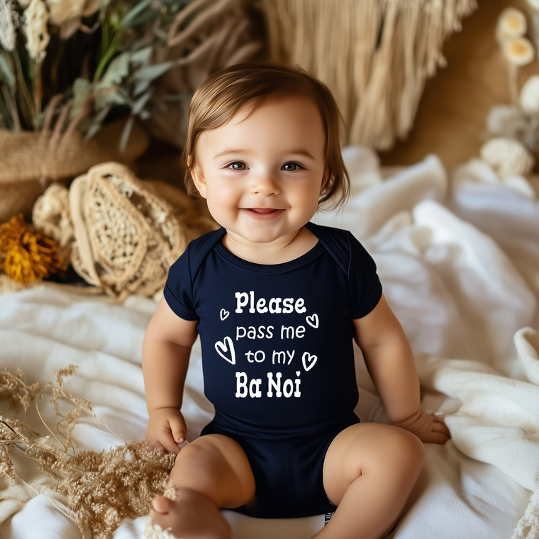 A smiling baby wearing a navy blue onesie with the text "Please pass me to my Ba Noi" is sitting on a bed with a floral background.