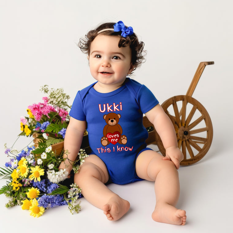 A smiling baby girl wearing a blue onesie with a teddy bear design and the text "Ukii loves me This I know" is sitting next to a wooden wagon filled with colorful flowers.