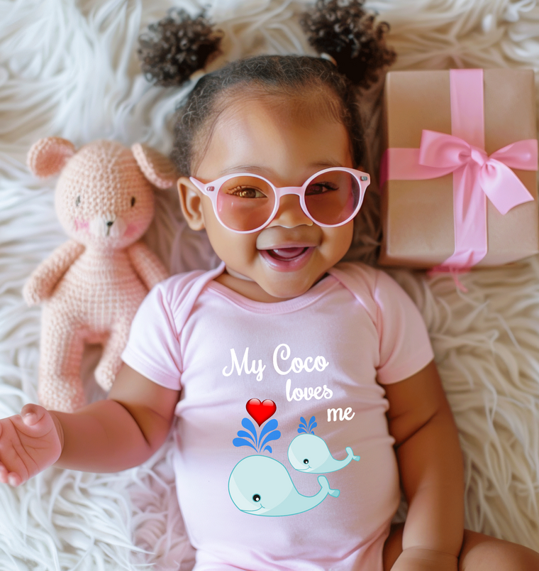 A smiling baby girl wearing pink glasses and a pink outfit with a whale graphic lies on a white fur rug next to a pink gift box.