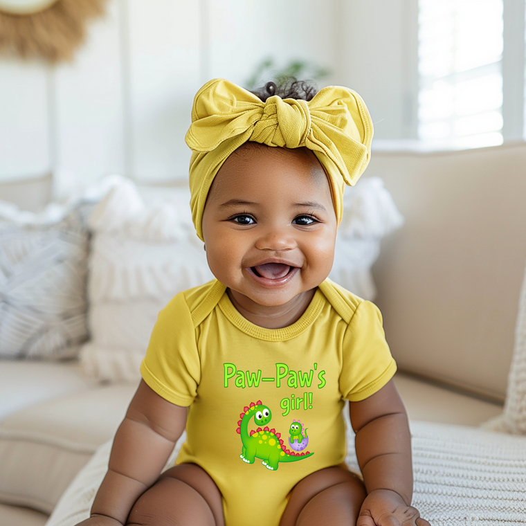 A smiling baby girl wearing a yellow outfit with a dinosaur graphic and the text "Paw-Paw's girl!" sits on a couch.