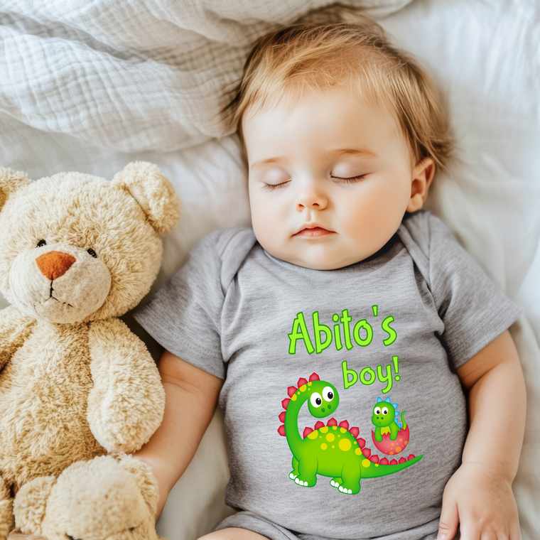 A baby sleeping next to a teddy bear, wearing a gray shirt with a dinosaur design and the text "Abito's boy!"