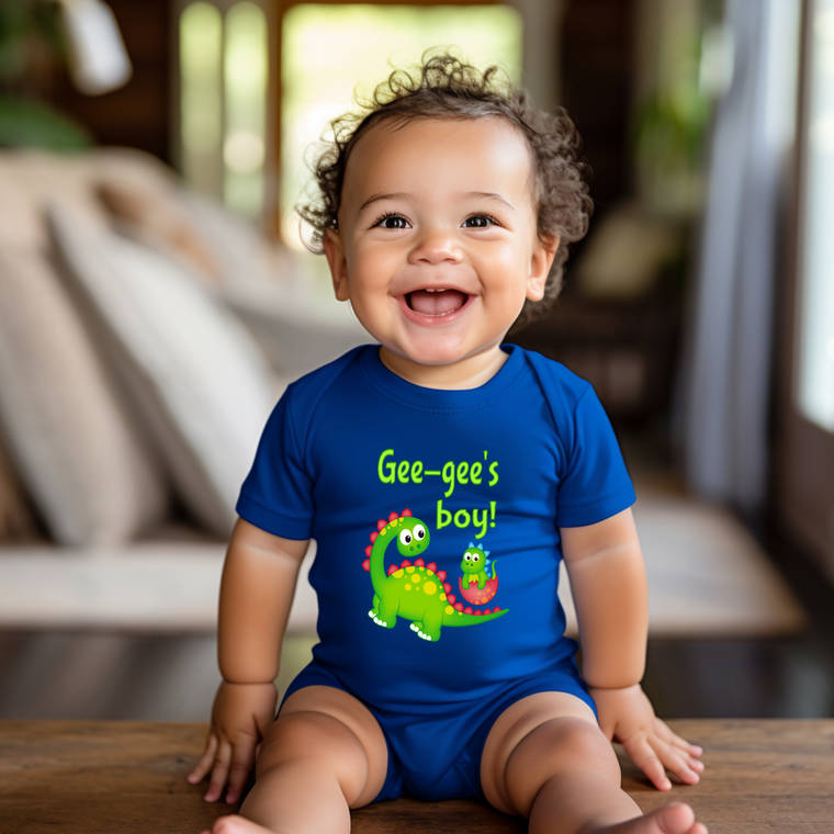 A smiling baby boy wearing a blue onesie with a dinosaur graphic and the text "Gee-gee's boy!"