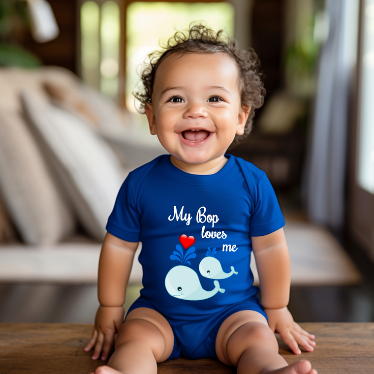 A smiling baby boy wearing a blue shirt with a whale graphic and the text "My Bop loves me" is sitting on a wooden surface.
