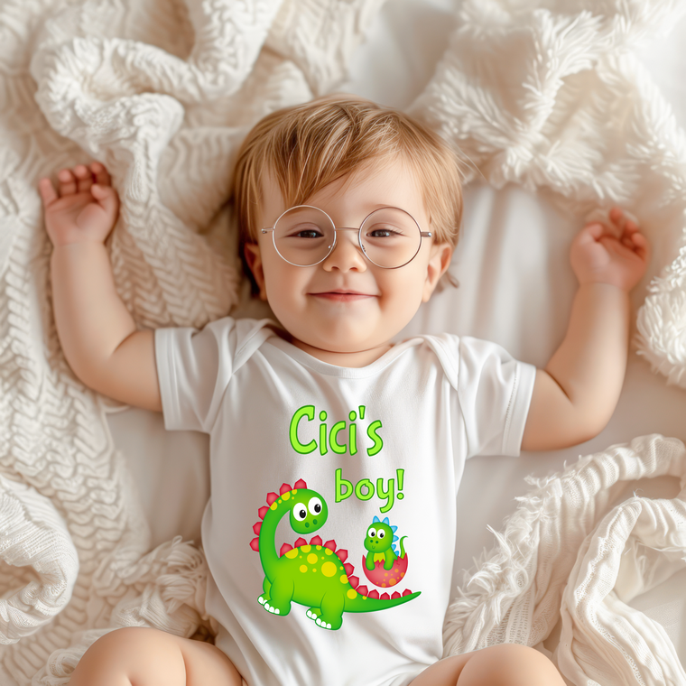 A smiling baby wearing a white onesie with a dinosaur graphic and the text "Cic's boy!" lying on a white blanket.