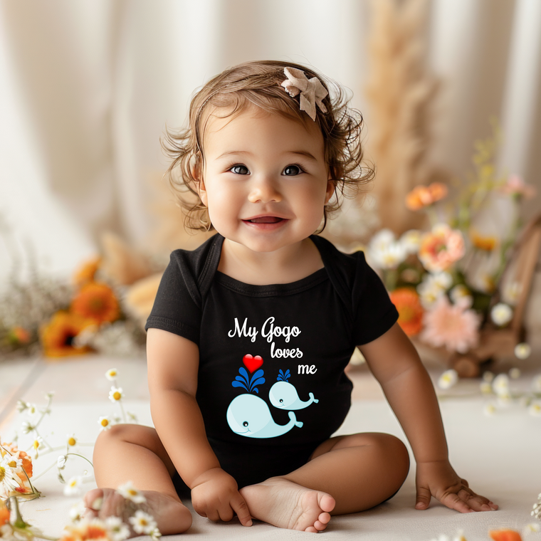 A smiling baby girl wearing a black shirt with a whale graphic and the text "My Gogo loves me" is sitting on the floor surrounded by flowers.