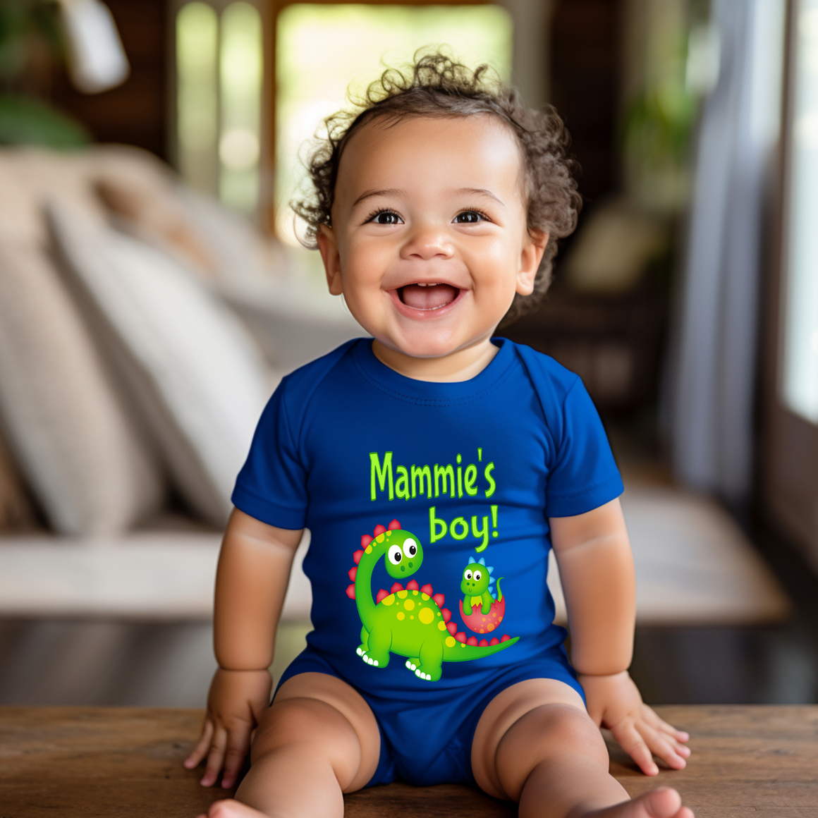 A smiling baby boy wearing a blue shirt with a dinosaur design and text that says "Mammie's boy!"
