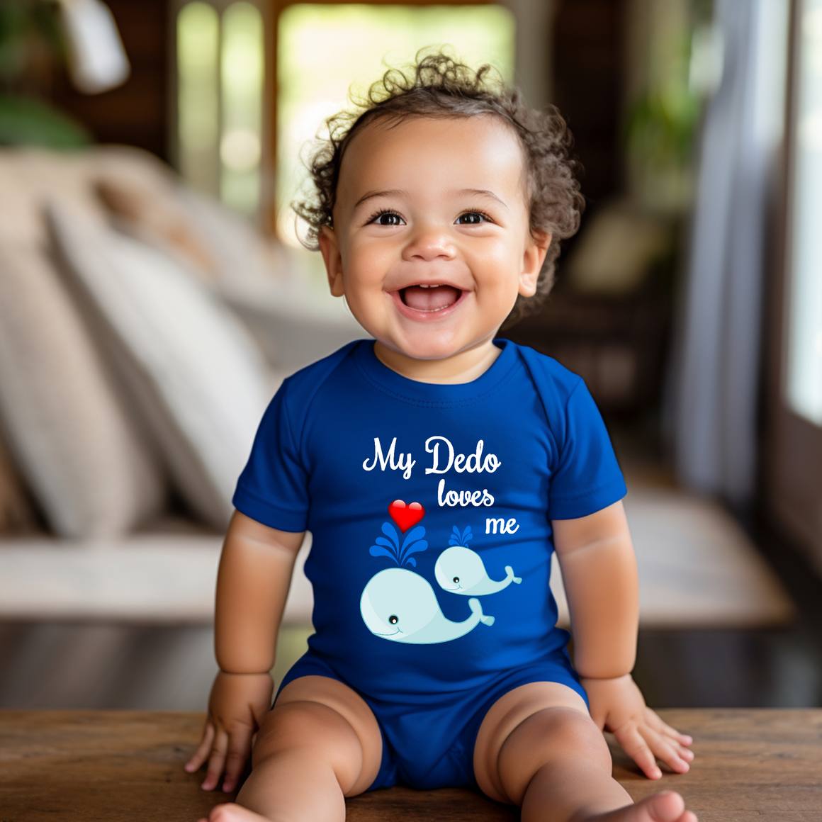A smiling baby boy wearing a blue shirt with a whale graphic and the text "My Dero loves me" is sitting on a wooden surface.
