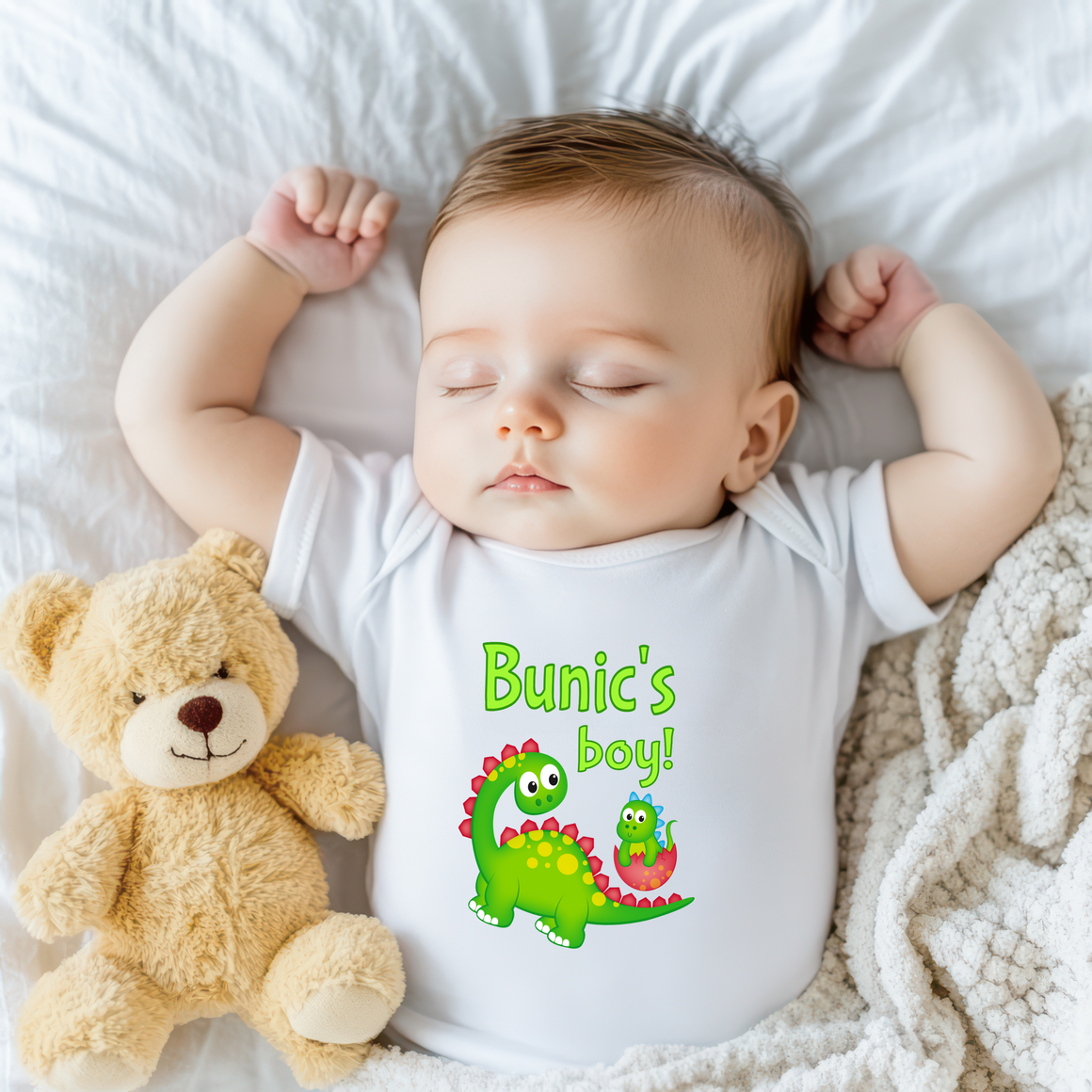 A sleeping baby wearing a white onesie with a dinosaur design and the text "Bunic's boy!" next to a teddy bear.