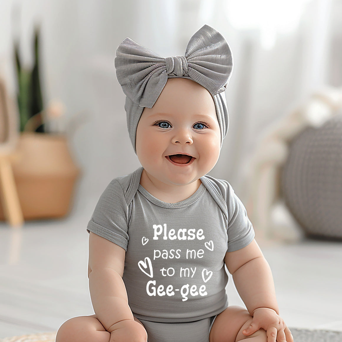 A smiling baby girl wearing a gray outfit with a bow and a t-shirt that says "Please pass me to my Gee-gee".
