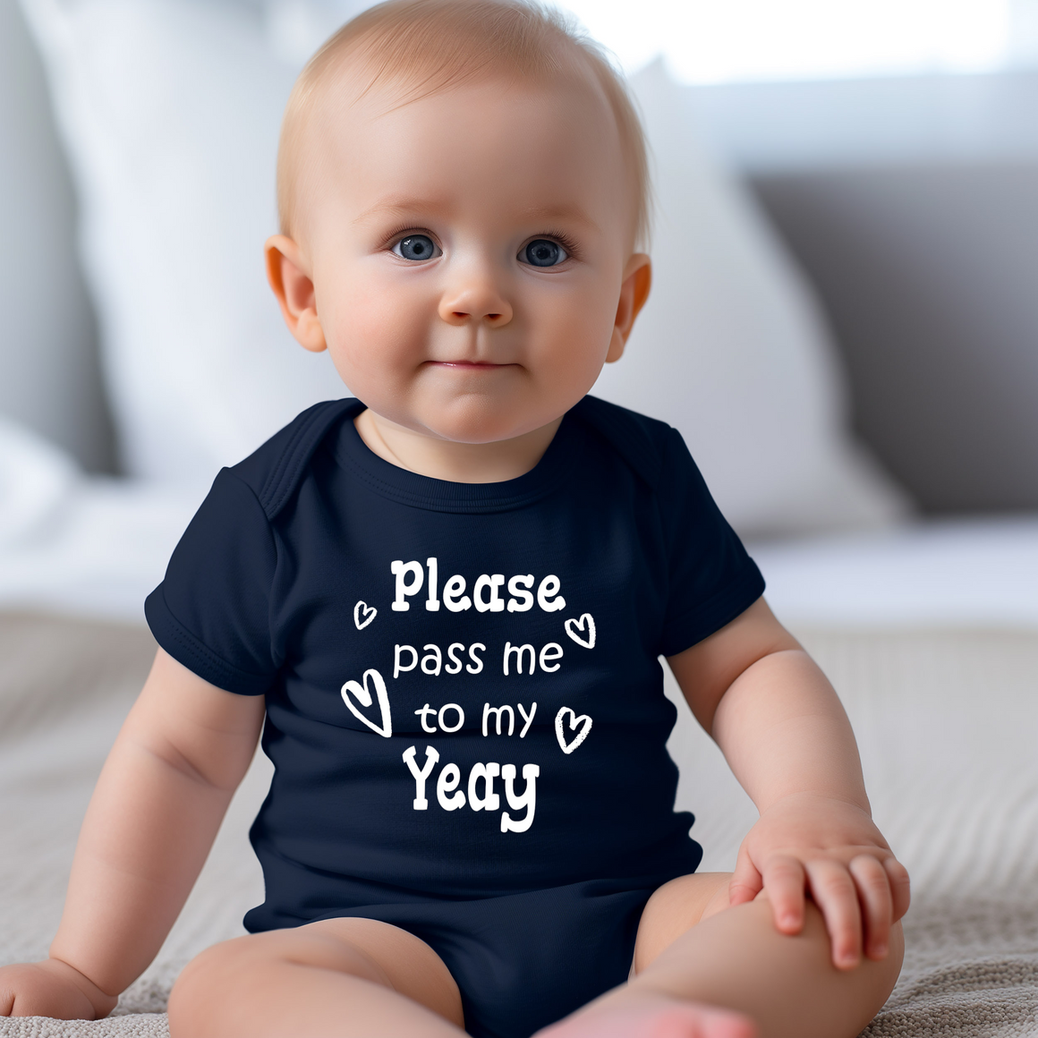 A smiling baby wearing a navy blue onesie with the text "Please pass me to my Yea" printed on it.