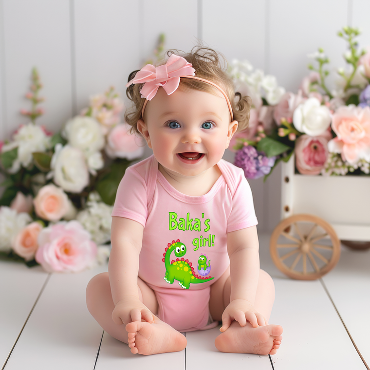 A smiling baby girl wearing a pink outfit with a dinosaur design sits on a wooden floor, surrounded by a bouquet of flowers.