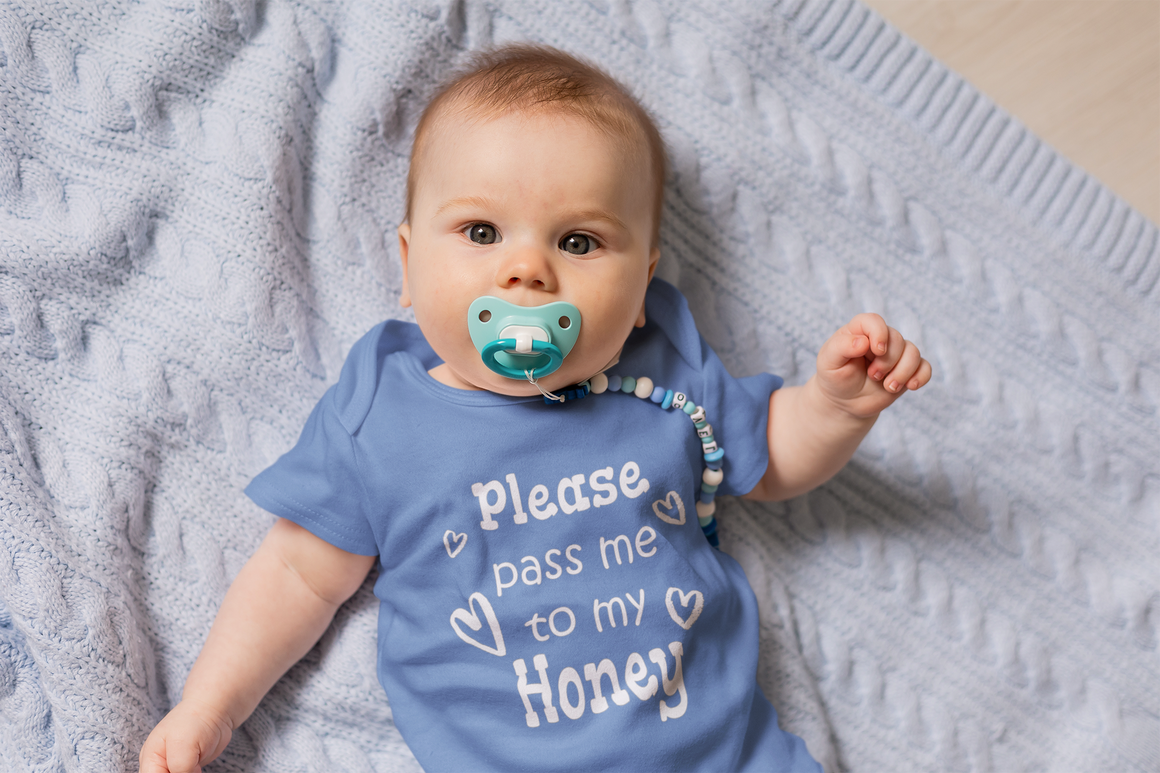 A baby wearing a blue onesie with the text "Please pass me to my Honey" lying on a blue blanket.